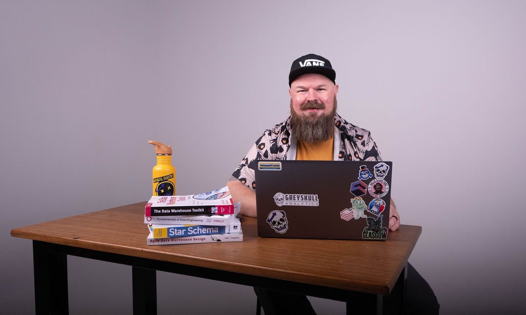 Johnny Winter at his desk with a laptop covered in Greyskull Analytics stickers and a stack of data engineering books
